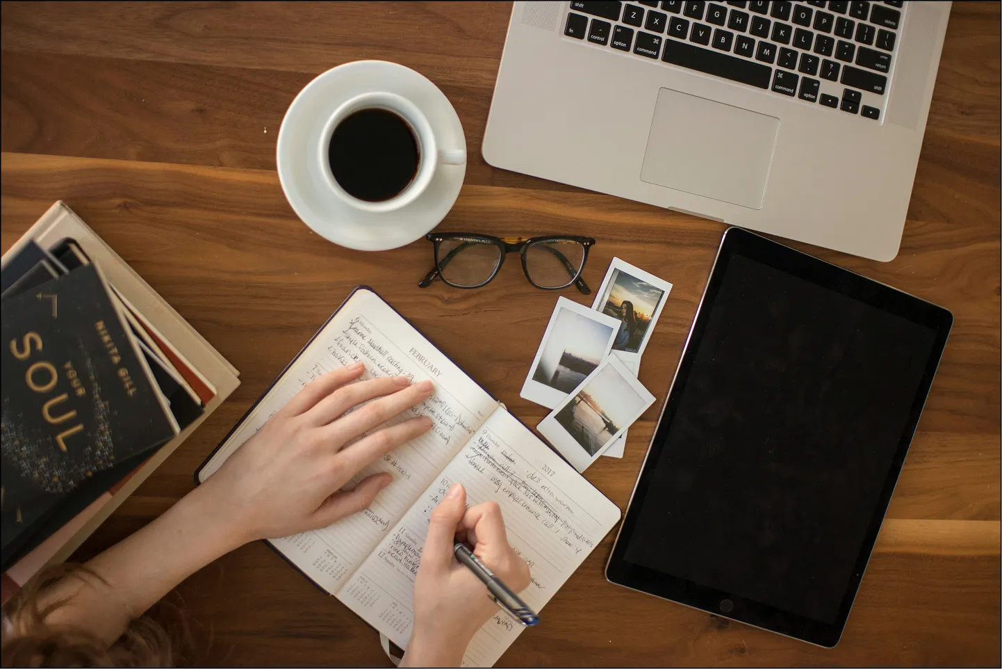 A person writing on a notebook next to a laptop and a cup of coffee.